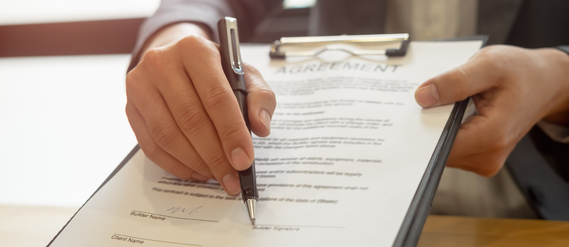 Individual Holding a clipboard and a pen with an agreement on the clipboard. The person is pointing the pen at the bottom, where the signature area is.